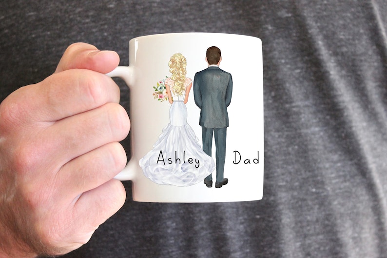 May include: White ceramic mug with a hand-drawn illustration of a bride and groom in wedding attire. The bride is holding a bouquet of flowers. The text "Ashley" and "Dad" is written below the couple.