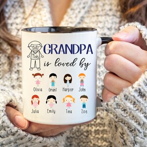 May include: White ceramic coffee mug with a black rim and handle. The mug has a blue inscription that reads "Grandpa is loved by" and features a cartoon drawing of a man and several children with their names below.