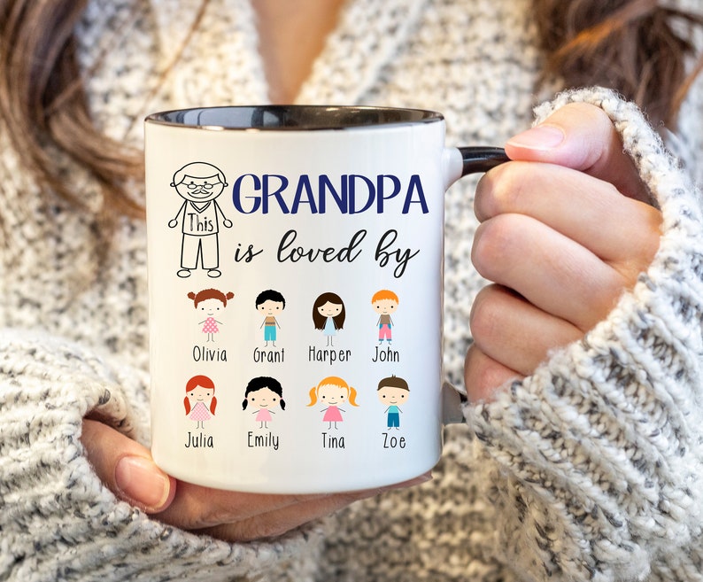 May include: White ceramic coffee mug with a black rim and handle. The mug has a blue inscription that reads "Grandpa is loved by" and features a cartoon drawing of a man and several children with their names below.