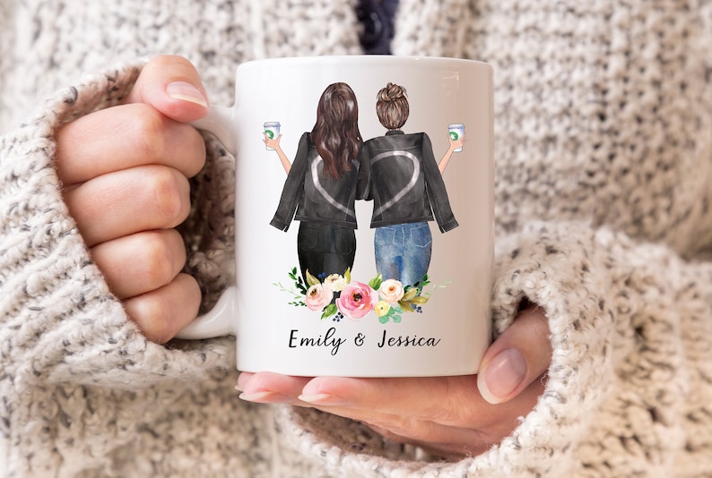 K&ouml;nnte beinhalten: Wei&szlig;e Keramiktasse mit einer Aquarell-Illustration von zwei Frauen in schwarzen Jacken, die Kaffeetassen halten. Die Frauen stehen R&uuml;cken an R&uuml;cken mit einem floralen Design am Boden der Tasse. Der Text "Emily & Jessica" ist unter dem Design gedruckt.