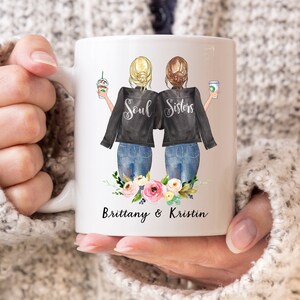 May include: White ceramic mug featuring a graphic of two women with blonde and brunette hair, wearing black jackets and jeans, holding coffee drinks. The text "Soul Sisters" is above a floral arrangement, with the names "Brittany & Kristin" below.