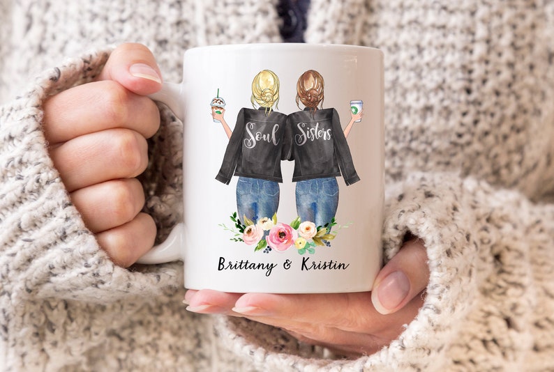 May include: White ceramic mug featuring a graphic of two women with blonde and brunette hair, wearing black jackets and jeans, holding coffee drinks. The text "Soul Sisters" is above a floral arrangement, with the names "Brittany & Kristin" below.