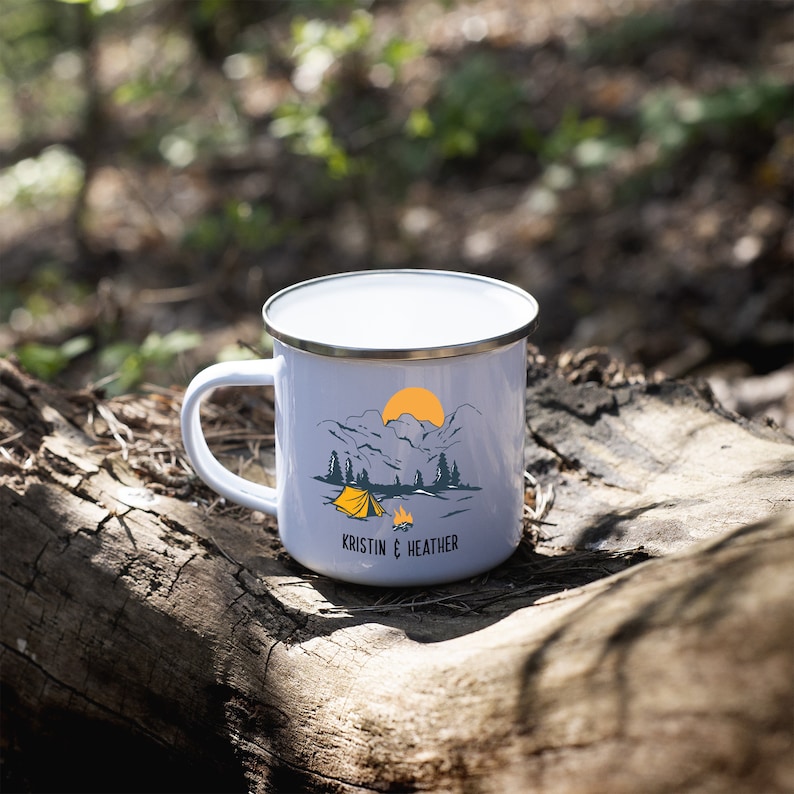 May include: White enamel camping mug with a mountain scene and a yellow tent. The mug has a silver rim and the text "Kristin & Heather" printed on the bottom.
