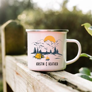 May include: White enamel camping mug with a mountain scene, a yellow tent, and a campfire. The mug has the text "KRISTIN & HEATHER" printed on the bottom.