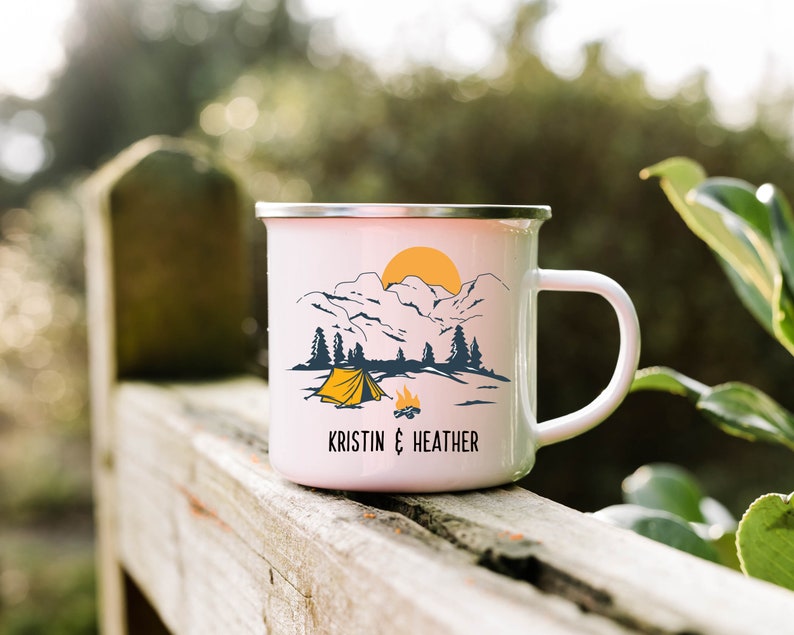 May include: White enamel camping mug with a mountain scene, a yellow tent, and a campfire. The mug has the text "KRISTIN & HEATHER" printed on the bottom.