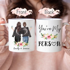 K&ouml;nnte beinhalten: Zwei wei&szlig;e Kaffeetassen mit einer personalisierten Botschaft. Die vordere Tasse zeigt eine Cartoon-Illustration von zwei Frauen in Jacken, die Kaffeetassen halten. Der Text auf der vorderen Tasse lautet "Emily & Jessica". Die hintere Tasse zeigt ein Blumenmuster und den Text "You're My Person" mit einem Herzen.