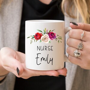 Puede incluir: Taza de cer&aacute;mica blanca con un dise&ntilde;o floral en tonos rojos, rosas y blancos. La taza dice "NURSE Emily" en texto negro.