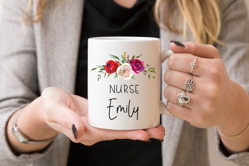 Puede incluir: Taza de cer&aacute;mica blanca con un dise&ntilde;o floral en tonos rojos, rosas y blancos. La taza dice "NURSE Emily" en texto negro.