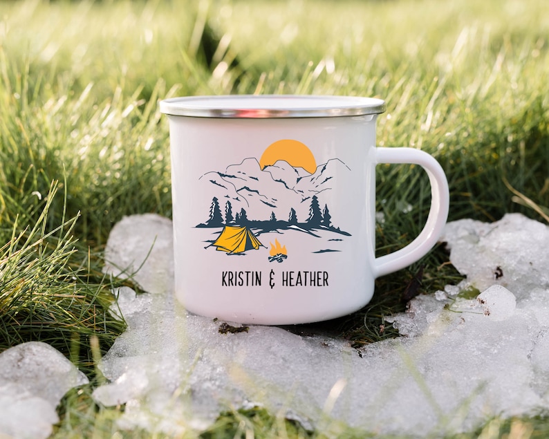 May include: White enamel camping mug with a mountain scene, a yellow tent, and a campfire. The mug has the text "KRISTIN & HEATHER" printed on the bottom.