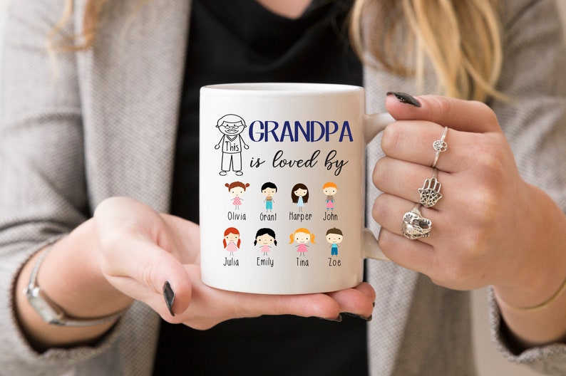 May include: White ceramic coffee mug with a black and white illustration of a grandpa and the text "Grandpa is loved by". Below the text are 8 stick figure illustrations of children with their names printed below each figure: Olivia, Grant, Harper, John, Julia, Emily, Tina, Zoe.
