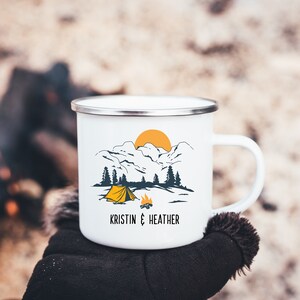 May include: White enamel camping mug with a mountain scene, a yellow tent, and a campfire. The mug has the text "Kristin & Heather" printed on the bottom.