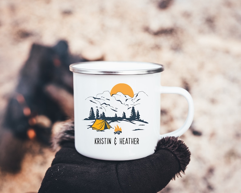 May include: White enamel camping mug with a mountain scene, a yellow tent, and a campfire. The mug has the text "Kristin & Heather" printed on the bottom.