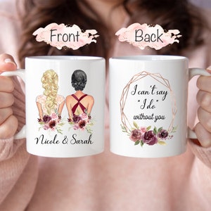 Puede incluir: Dos tazas de cerámica blanca con un diseño floral. La taza delantera tiene un dibujo de dos mujeres con atuendo de boda con los nombres "Nicole & Sarah" impresos debajo. La taza trasera tiene un marco geométrico dorado con el texto "No puedo decir "Sí, quiero" sin ti" en su interior.
