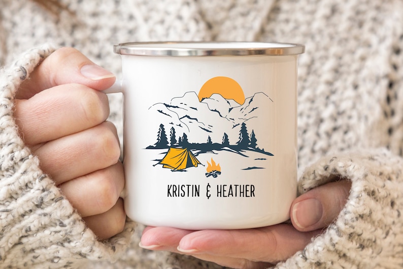May include: White enamel camping mug with a mountain scene, a yellow tent, a campfire, and a yellow sun. The mug has the text "KRISTIN & HEATHER" printed on the bottom.