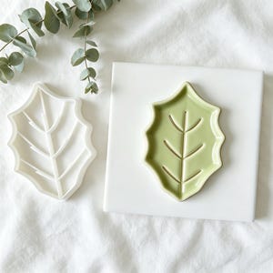May include: A white plastic leaf-shaped cookie cutter and a matching light green ceramic leaf-shaped dish with a central vein design. The dish is placed on a square white tile, and a sprig of eucalyptus is positioned above the items.