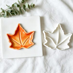 May include: A glossy orange ceramic leaf-shaped dish rests on a square white plate next to a white plastic leaf-shaped cookie cutter. The dish features embossed vein details and a smooth, curved edge, mirroring the shape of the white cutter.