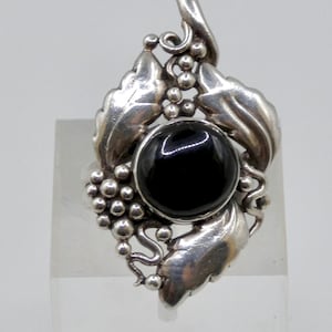 May include: A silver-toned ring featuring a large, round black stone at its center. The ring is adorned with detailed leaf and grape cluster designs, creating a nature-inspired aesthetic. The ring is set against a neutral background.