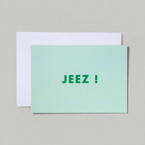 May include: A mint green greetings card with the word "JEEZ!" printed in dark green. A white envelope is behind the card. The card and envelope are on a light grey surface.