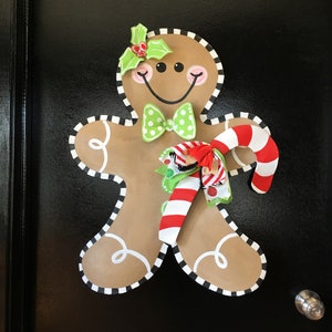 May include: A gingerbread person wall decoration featuring a tan body with white icing swirls, a black and white striped border, a green polka dot bow tie, a holly sprig on the head, and a red and white candy cane held with a multi-colored ribbon.