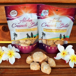 May include: Two bags of Aloha Crunch Bites dog treats, one with coconut and one with honey, on a wooden surface with two white frangipani flowers. The bags are labelled "Aloha Crunch Bites" and "Bite-sized Aloha for your dog".