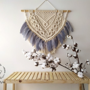 Può includere: Applique da parete in macramè color crema con un design geometrico, impreziosito da piume grigie e perline di legno. La decorazione murale è sospesa a un'asta di legno. Un rametto di cotone aggiunge un tocco naturale.