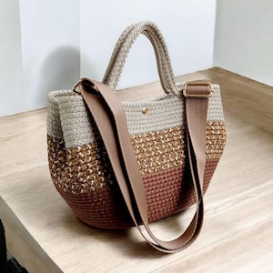 K&ouml;nnte beinhalten: Eine braune, beige und wei&szlig;e gestreifte geh&auml;kelte Tragetasche mit einem braunen Lederriemen. Die Tasche hat einen abgerundeten Boden und zwei obere Henkel.