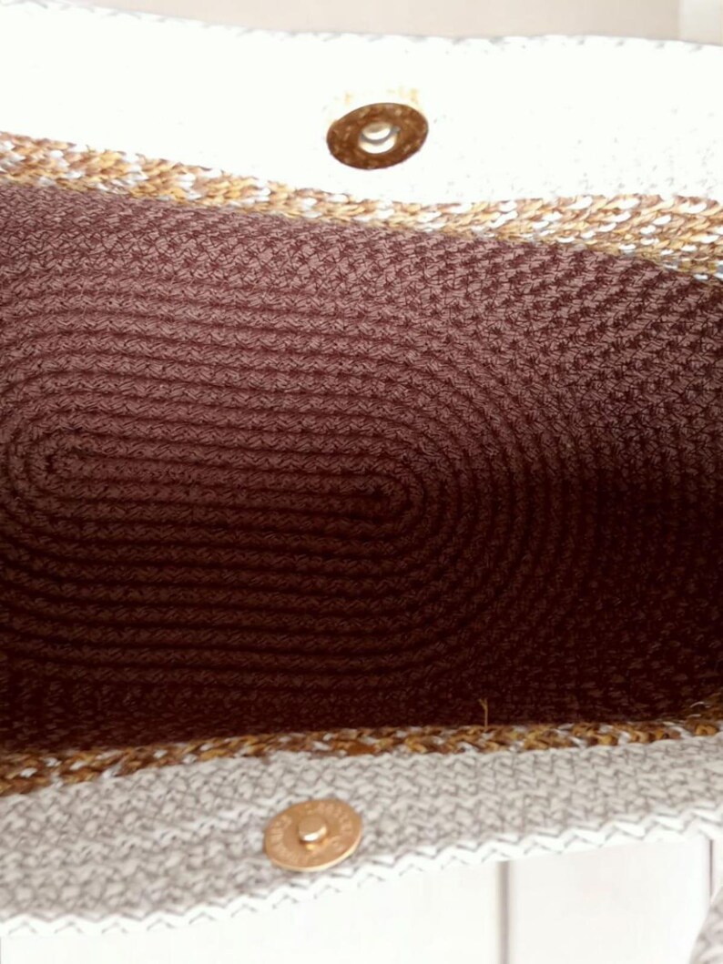 K&ouml;nnte beinhalten: Nahaufnahme des Inneren einer wei&szlig;en und braunen gewebten Tragetasche. Die Tasche hat ein braunes gewebtes Futter und einen goldenen Metallknopf.