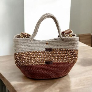 K&ouml;nnte beinhalten: Eine geh&auml;kelte Tragetasche in Braun, Beige und Wei&szlig; mit Ledergriffen. Die Tasche hat einen abgerundeten Boden und eine gro&szlig;e &Ouml;ffnung.