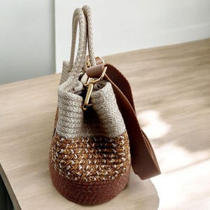 K&ouml;nnte beinhalten: Eine gewebte Tragetasche in Braun und Creme mit einem braunen Lederriemen. Die Tasche hat einen oberen Griff und einen langen Schulterriemen.