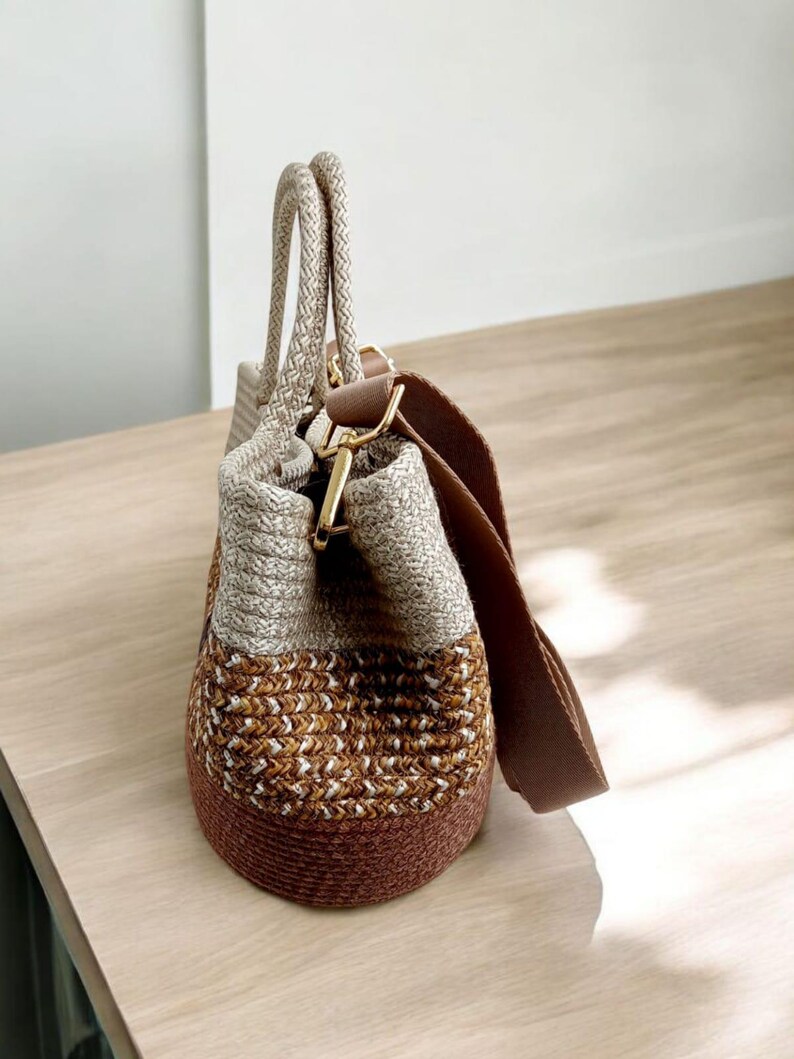 K&ouml;nnte beinhalten: Eine gewebte Tragetasche in Braun und Creme mit einem braunen Lederriemen. Die Tasche hat einen oberen Griff und einen langen Schulterriemen.