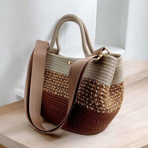 K&ouml;nnte beinhalten: Eine braune, beige und wei&szlig;e gestreifte geh&auml;kelte Tragetasche mit einem braunen Lederriemen. Die Tasche hat einen oberen Griff und einen langen Schulterriemen.