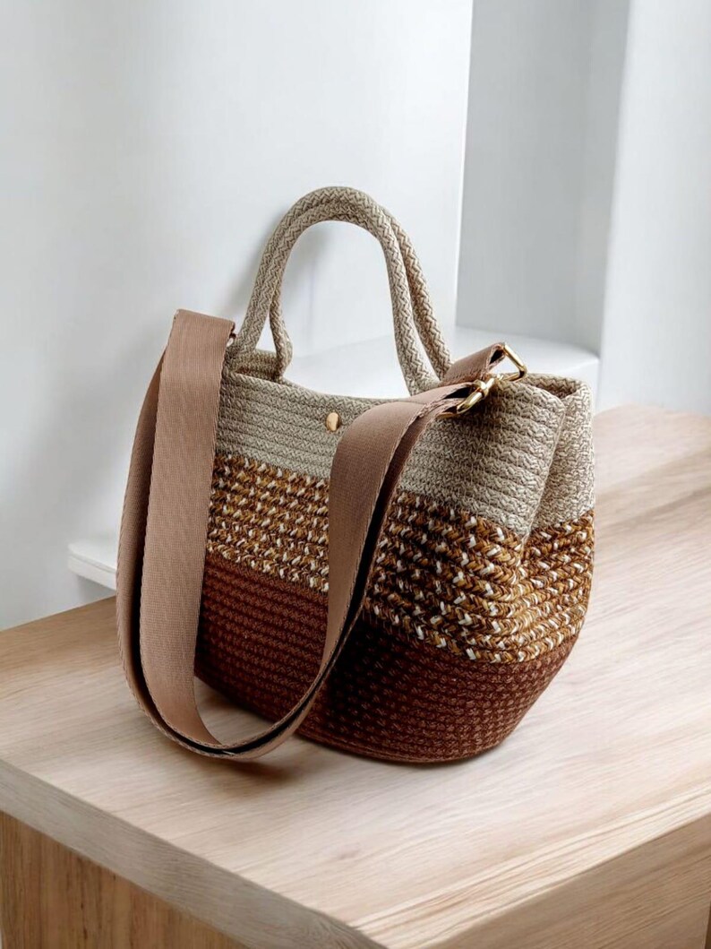 K&ouml;nnte beinhalten: Eine braune, beige und wei&szlig;e gestreifte geh&auml;kelte Tragetasche mit einem braunen Lederriemen. Die Tasche hat einen oberen Griff und einen langen Schulterriemen.