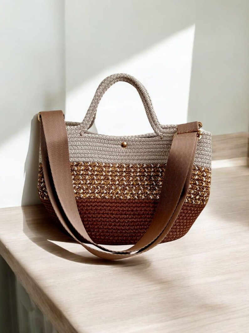 K&ouml;nnte beinhalten: Eine braune, beige und wei&szlig;e geh&auml;kelte Tragetasche mit einem braunen Lederriemen. Die Tasche hat einen oberen Griff und einen langen Riemen, der &uuml;ber der Schulter getragen werden kann.