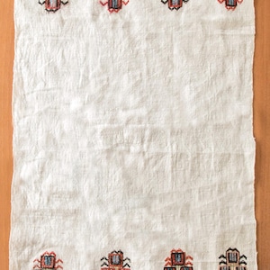 May include: White linen textile with embroidered geometric patterns in red, blue, and brown. The top and bottom borders feature repeating stylized motifs. The fabric has a slightly wrinkled texture.