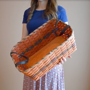 May include: A woven basket with brown and orange stripes and leather handles. The basket is empty and has a light brown lining.