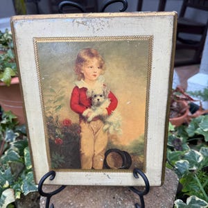 May include: Vintage framed print of a young child in a red jacket and tan pants, holding a fluffy white dog. The print is set in a cream-colored frame with gold trim. A black hat rests at the child's feet. The artwork is displayed on a black metal stand.