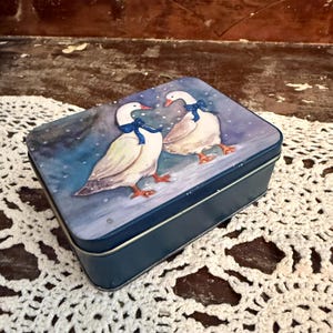 May include: A rectangular, blue metal tin with a painted illustration of two white geese wearing blue bows. The geese stand on a snowy surface against a blue and purple background. The tin is on a white, lace-like surface.