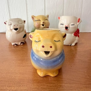 May include: A collection of four ceramic pig-shaped planters or containers. One yellow pig with a blue band sits in the foreground. Behind it are three other ceramic pigs: one white with black spots, one solid yellow, and one white with a red bow.