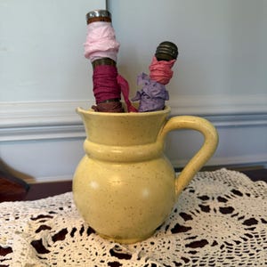 May include: A yellow ceramic pitcher with a handle, speckled with brown dots, holds two objects wrapped in pink, purple, and burgundy fabric. The pitcher sits on a white lace doily, creating a rustic, vintage aesthetic.