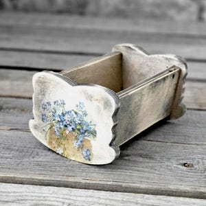 Könnte beinhalten: Kleine, rustikale Holzkiste mit floralem Design. Die Kiste ist in Grau- und Weißtönen bemalt und hat auf der Vorderseite eine Dekoration aus blauen Blumen. Die Kiste hat einen gewellten Rand und ist für kleine Gegenstände konzipiert.