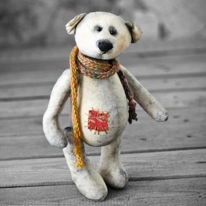 Könnte beinhalten: Ein handgefertigter, cremefarbener Teddybär mit einem bunten Schal und einem roten Flicken auf dem Bauch. Der Bär hat schwarze Knopfaugen und eine schwarze Nase. Er steht auf einer Holzoberfläche.