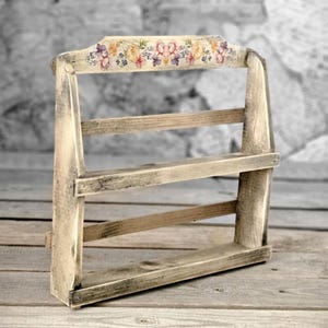 Op de afbeelding: Een rustiek houten plank met twee niveaus, geschilderd in een verweerde witte afwerking. De bovenste plank heeft een decoratief bloemenontwerp in tinten roze, paars en geel.