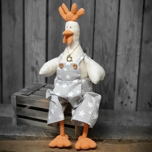 Pode incluir: Um frango de pelúcia branco vestindo um macacão cinza com estrelas brancas e um colar de ouro. O frango está sentado em uma caixa de madeira.