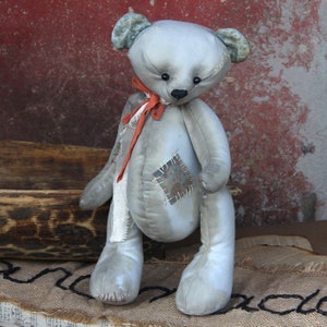 Könnte beinhalten: Ein grauer, abgenutzter Teddybär mit einem roten Band um den Hals. Der Bär hat einen Flicken auf seiner Brust und sitzt auf einer Jutestruktur.