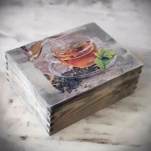 Op de afbeelding: Een houten doos met een scharnierend deksel, met een bedrukte afbeelding van een glazen theekopje met een theezakje en een takje munt. De doos heeft een verweerde, rustieke uitstraling met een lichtgrijze en bruine afwerking.