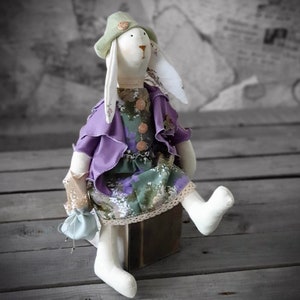 May include: A white fabric bunny doll wearing a purple jacket with a floral print dress and a green hat. The doll is sitting on a wooden box.