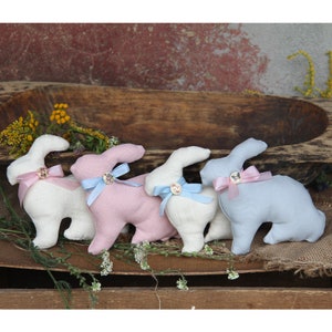 Può includere: Quattro coniglietti di stoffa fatti a mano in bianco, rosa e blu con dettagli in nastro. I coniglietti sono seduti su una superficie di legno.