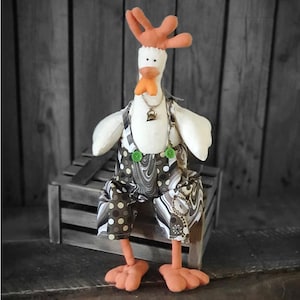Pode incluir: Um boneco de frango de tecido branco vestindo um macacão estampado em marrom e branco com botões verdes e um colar de corrente dourada com um sino. O frango está sentado em uma caixa de madeira.