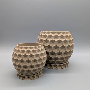 Puede incluir: Dos macetas beige con patrones geométricos de diferentes tamaños. Las macetas tienen forma esférica con un patrón texturizado en forma de diamante. Cada maceta se asienta sobre una base de platillo a juego. El fondo es gris claro liso.