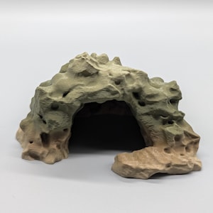 Op de afbeelding: Een getextureerde, beige en lichtbruine rotsvormige grottenstructuur met een holle binnenkant en een boogvormige opening. Het buitenoppervlak vertoont onregelmatige bulten, spleten en kleine gaatjes die het uiterlijk van een natuurlijke steen nabootsen.